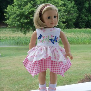 May include: A doll wearing a pink and white dress with a floral and butterfly design. The doll has blonde hair and blue eyes and is wearing pink shoes.