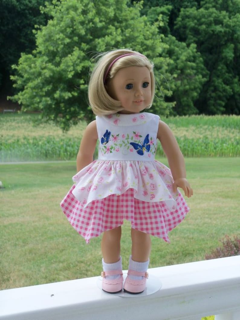 May include: A doll wearing a pink and white dress with a floral and butterfly design. The doll has blonde hair and blue eyes and is wearing pink shoes.