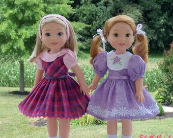 May include: Two 14-inch Wellie Wishers dolls wearing dresses. The doll on the left is wearing a pink and purple plaid dress with a pink collar and white trim. The doll on the right is wearing a lavender dress with white embroidered flowers and a white collar. The dolls are standing on a white surface with the text "Wellie Best Friends" in purple.