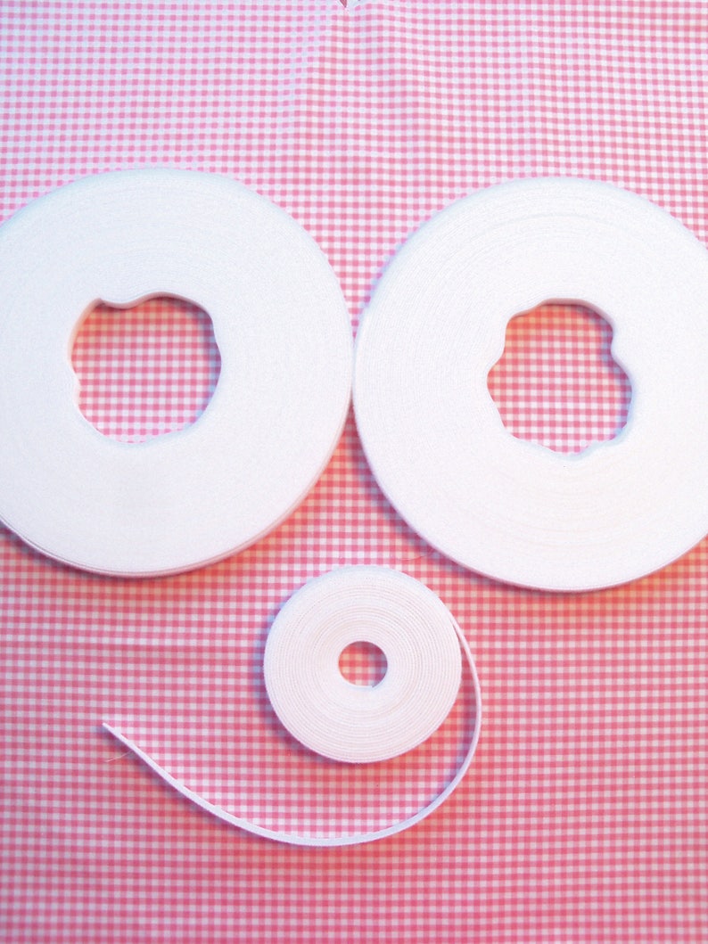 May include: Three spools of white elastic ribbon are arranged on a pink and white gingham background. Two large spools are side by side, and a smaller spool is below them. The ribbon is tightly wound.