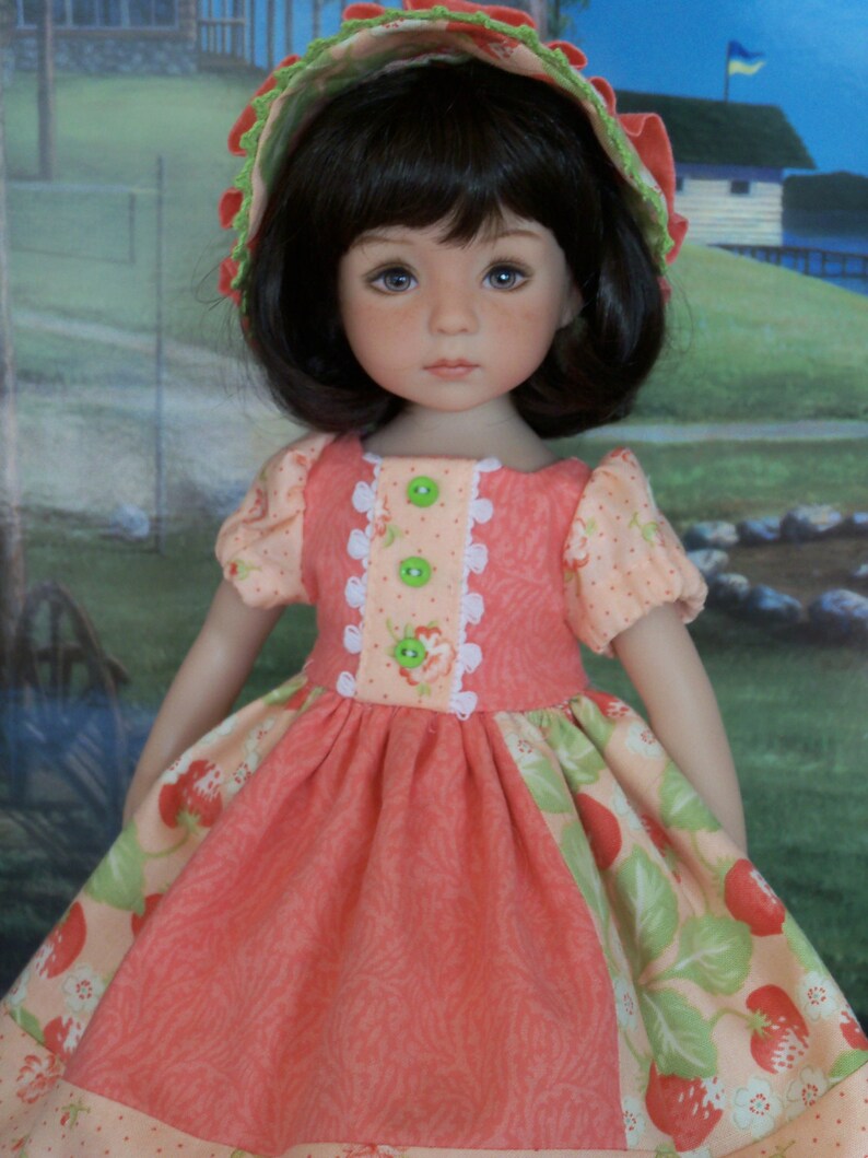 May include: A doll wearing a pink and green dress with a strawberry print and a matching green and white hat. The doll has brown hair and blue eyes.