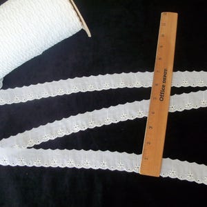 May include: White eyelet lace trim with a scalloped edge and small floral cutouts. A wooden ruler, marked in inches, is placed across the trim. A roll of the same lace is visible in the background. The trim is approximately 1 inch wide.