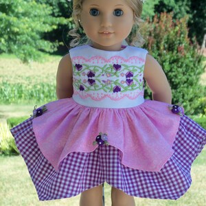 May include: A doll wearing a pink and white dress with a purple gingham skirt. The doll has blonde hair and blue eyes and is wearing a flower crown.