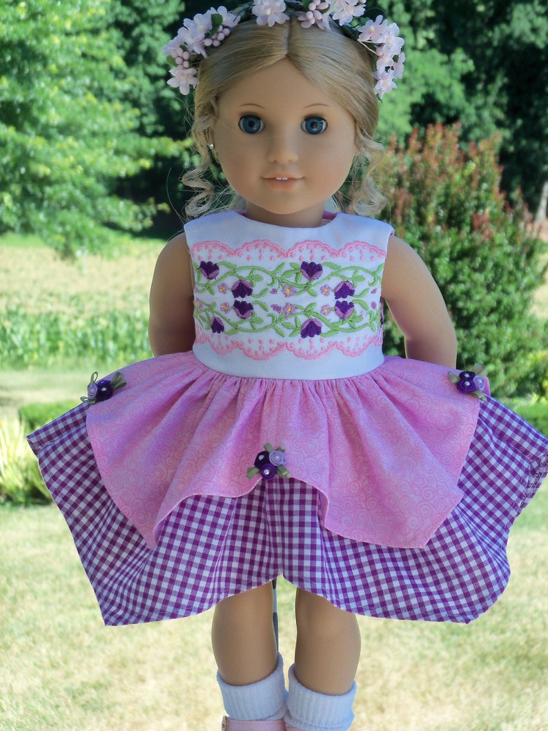 May include: A doll wearing a pink and white dress with a purple gingham skirt. The doll has blonde hair and blue eyes and is wearing a flower crown.