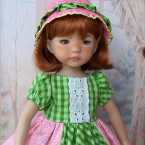 May include: A doll wearing a green and pink gingham dress with a white lace trim and a pink and green hat. The doll has red hair and blue eyes.