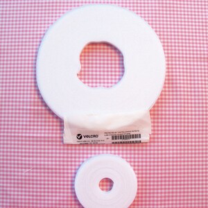 May include: Two rolls of white Velcro hook and loop tape, one large and one small, with a central hole. A small white package with the Velcro logo and text is between the rolls. The background is a pink and white gingham pattern.