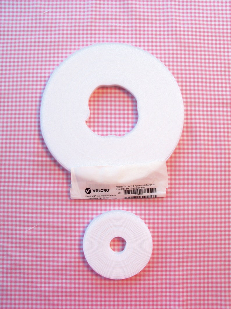 May include: Two rolls of white Velcro hook and loop tape, one large and one small, with a central hole. A small white package with the Velcro logo and text is between the rolls. The background is a pink and white gingham pattern.