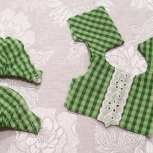 May include: Green and white gingham fabric pieces with white lace trim. The fabric is cut into different shapes for sewing a garment.