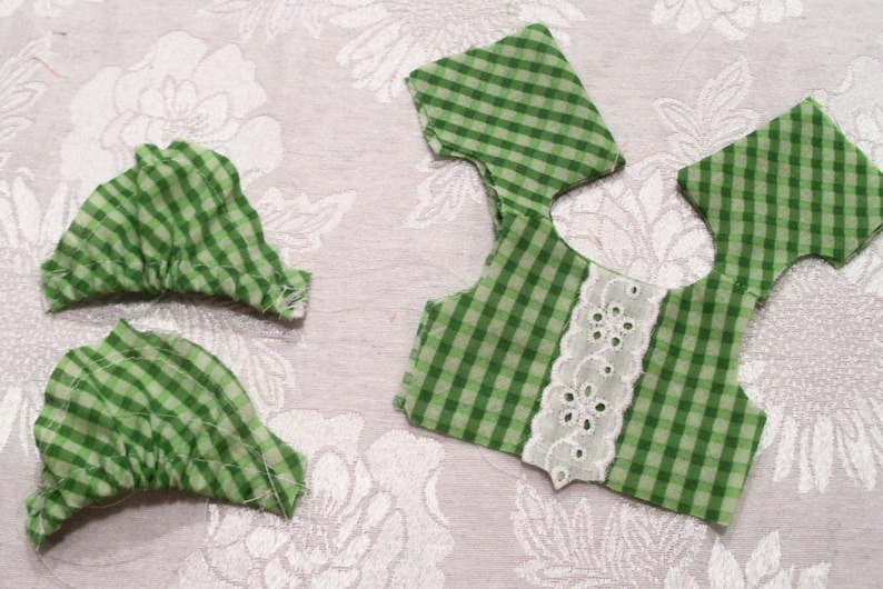 May include: Green and white gingham fabric pieces with white lace trim. The fabric is cut into different shapes for sewing a garment.