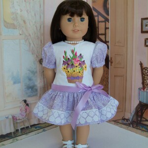 May include: A doll wearing a lavender dress with a white lace trim and a floral embroidered design. The dress has a lavender satin ribbon tied around the waist. The doll is wearing white sandals.