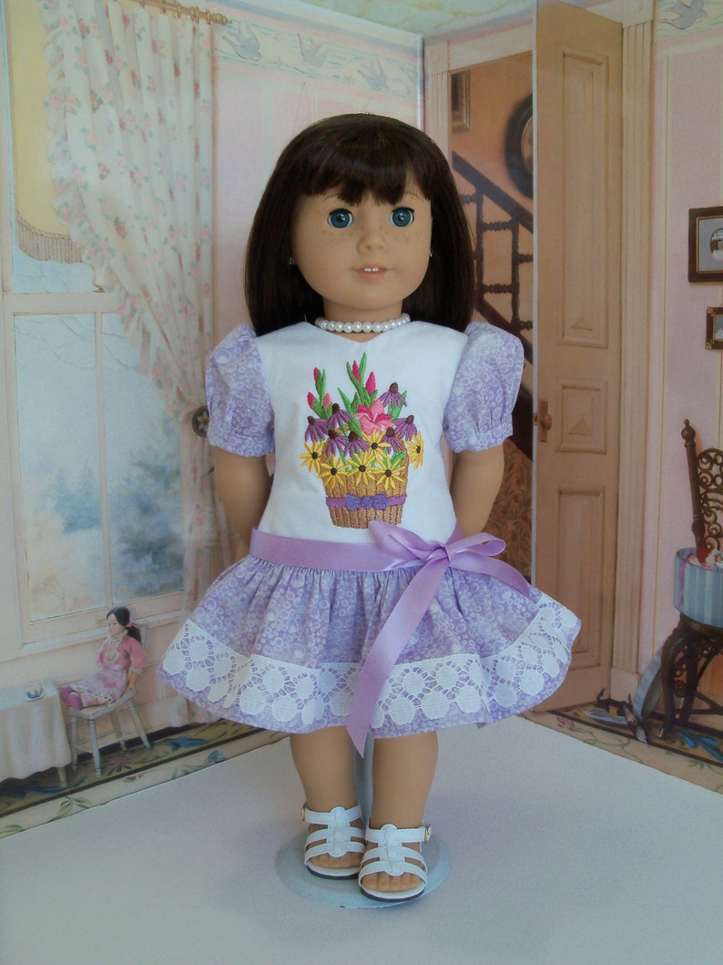 May include: A doll wearing a lavender dress with a white lace trim and a floral embroidered design. The dress has a lavender satin ribbon tied around the waist. The doll is wearing white sandals.