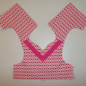 May include: Pink fabric with a diamond pattern, cut out for a garment with a pink trim along the neckline.