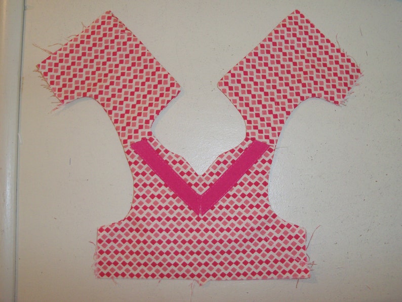 May include: Pink fabric with a diamond pattern, cut out for a garment with a pink trim along the neckline.
