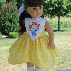 May include: A yellow dress with a white embroidered floral design on the bodice. The dress has a white collar and a white bow. The doll is wearing white socks and white shoes.