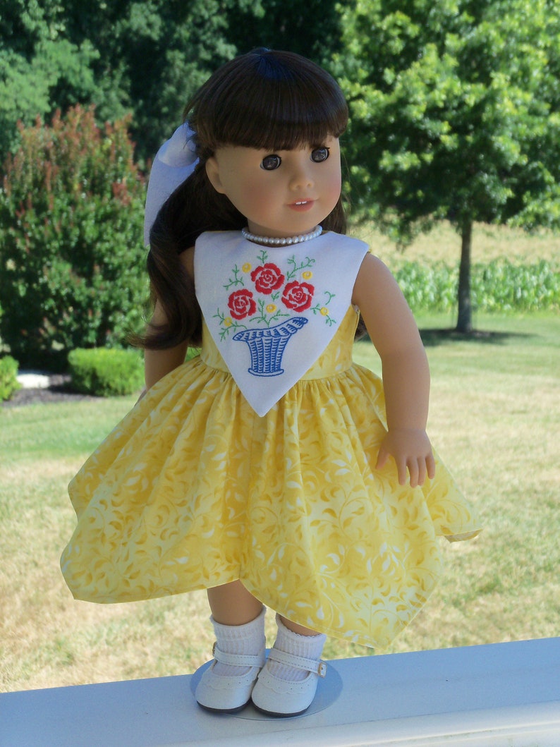May include: A yellow dress with a white embroidered floral design on the bodice. The dress has a white collar and a white bow. The doll is wearing white socks and white shoes.