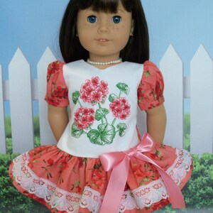 May include: A doll wearing a white dress with a pink bow and a floral print. The dress has a lace trim and the doll has brown hair and blue eyes.