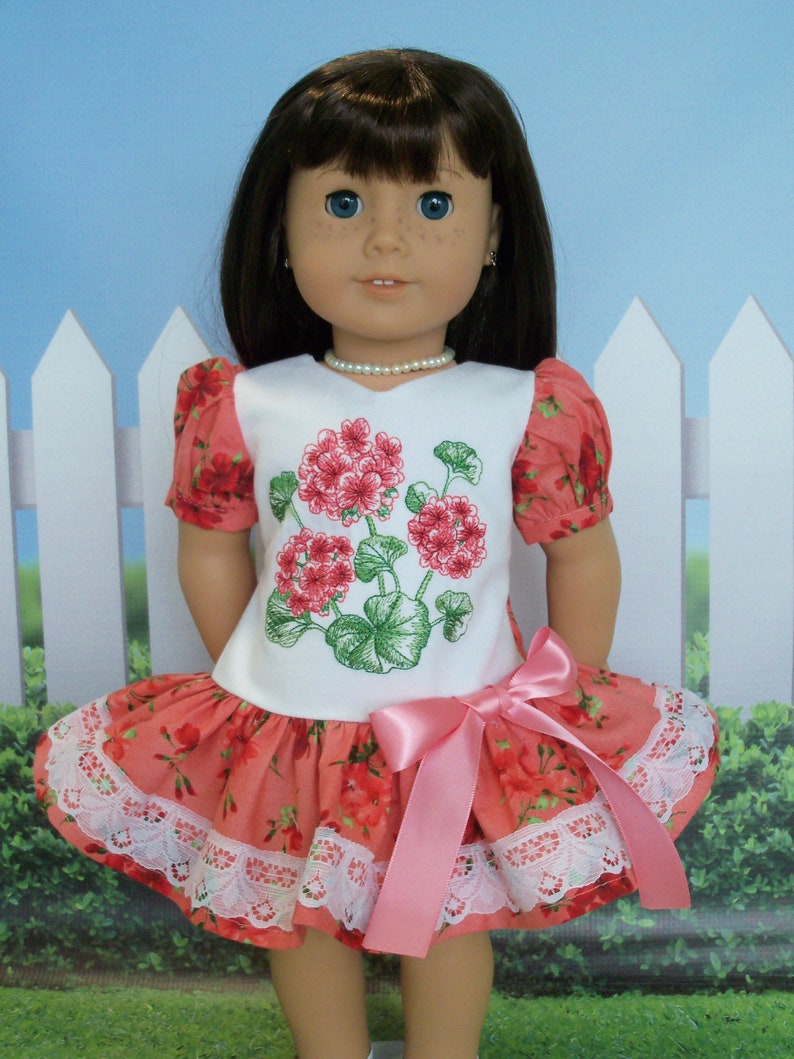 May include: A doll wearing a white dress with a pink bow and a floral print. The dress has a lace trim and the doll has brown hair and blue eyes.