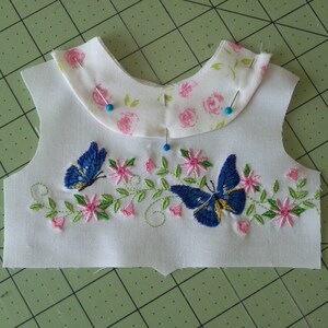 May include: A white fabric with a floral pattern and two blue butterflies embroidered on it. The fabric is pinned to a green cutting mat.