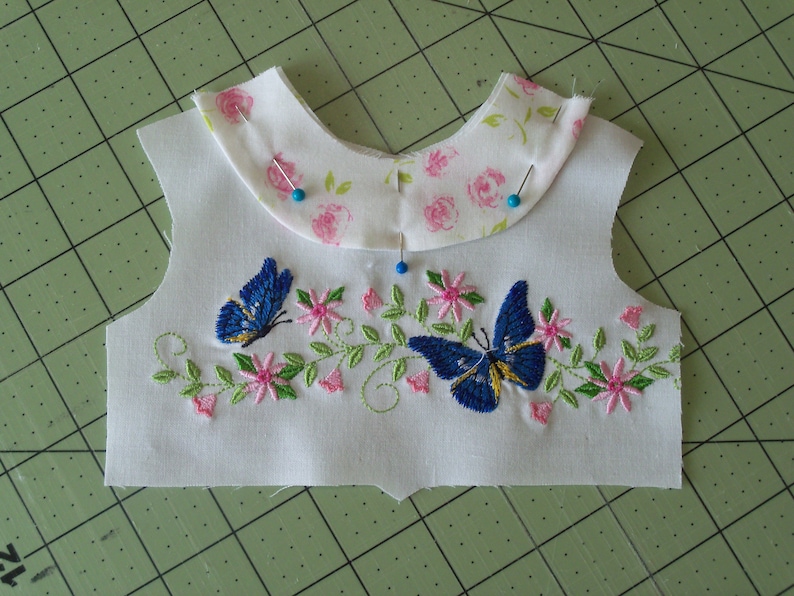 May include: A white fabric with a floral pattern and two blue butterflies embroidered on it. The fabric is pinned to a green cutting mat.