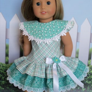 May include: A doll wearing a green and white plaid dress with a white lace trim and a white satin bow. The dress has a green and white floral print on the bottom layer. The doll has blonde hair and blue eyes.