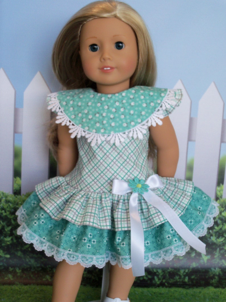 May include: A doll wearing a green and white plaid dress with a white lace trim and a white satin bow. The dress has a green and white floral print on the bottom layer. The doll has blonde hair and blue eyes.