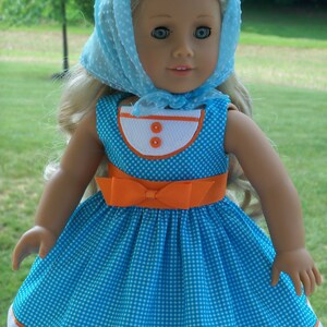 May include: A doll wearing a blue and white gingham dress with an orange sash and a light blue dotted scarf. The doll has blonde hair and blue eyes.