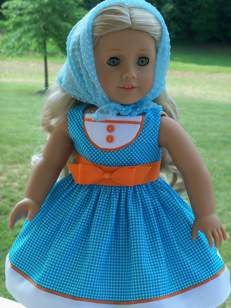 May include: A doll wearing a blue and white gingham dress with an orange sash and a light blue dotted scarf. The doll has blonde hair and blue eyes.