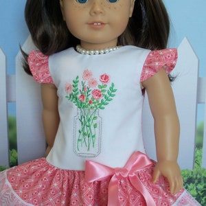 May include: A white dress with pink and white checkered fabric and a lace trim. The dress has a pink satin bow and features an embroidered design of a jar of flowers.