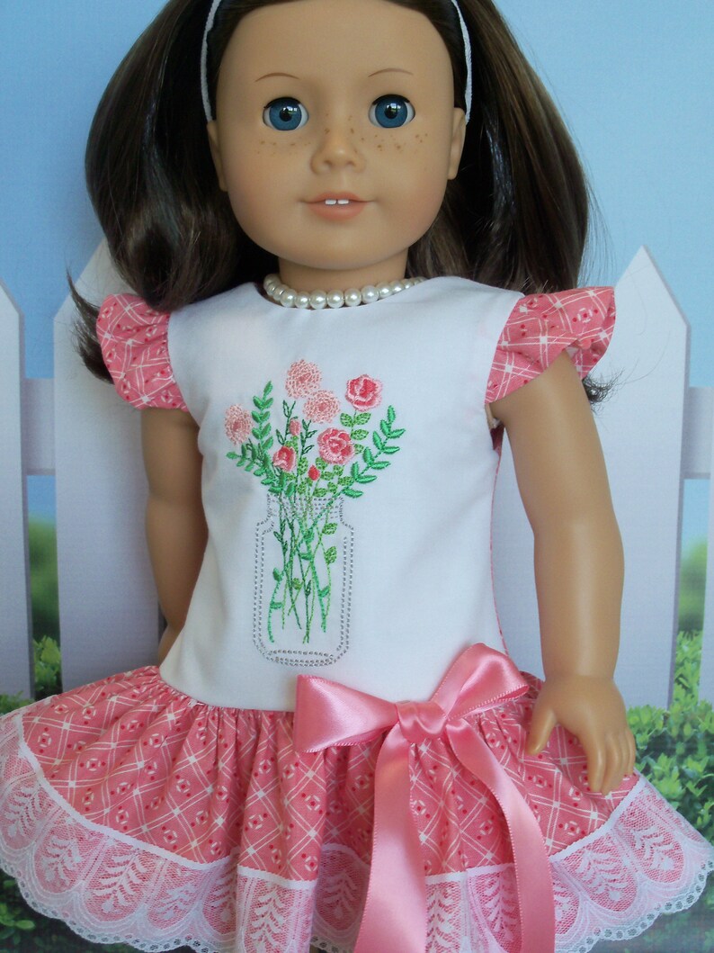 May include: A white dress with pink and white checkered fabric and a lace trim. The dress has a pink satin bow and features an embroidered design of a jar of flowers.