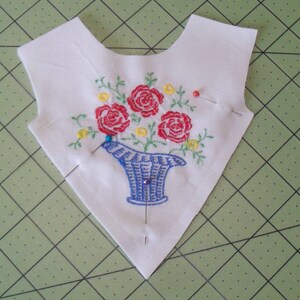 May include: A white fabric triangle with a blue and white embroidered flower arrangement. The flowers are red, yellow, and green. There are several pins holding the fabric in place.