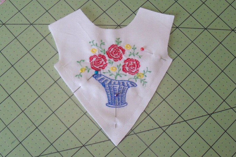 May include: A white fabric triangle with a blue and white embroidered flower arrangement. The flowers are red, yellow, and green. There are several pins holding the fabric in place.