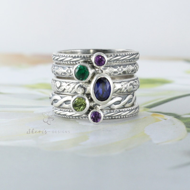 Set of 5 silver rings with birthstones stacked together. Top and bottom ring bands hold round 3mm gems. The next two bands hold round 4mm gems. The middle band holds a 6x4mm oval band. All bands are patterned and show the oxidized finish.