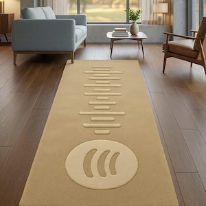 Puede incluir: Una alfombra rectangular beige con un diseño crema que se asemeja a una forma de onda musical y un logotipo circular. La alfombra está colocada sobre un suelo de madera oscura en un salón. Las dimensiones de la alfombra no son visibles.