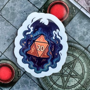 Può includere: Un adesivo raffigurante un dado D20 con il numero 20, circondato da fumo blu e una sagoma di drago. Il dado è arancione con dettagli dorati. L'adesivo è su una superficie grigia con altri elementi di gioco.