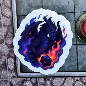 Könnte beinhalten: Ein Aufkleber mit einem dunkelblauen Drachen mit roten Augen, umgeben von lila und orangefarbenen Flammen. Der Drache hält einen schwarzen und grauen Würfel. Der Aufkleber hat einen weißen Rand und liegt auf einer grauen Oberfläche.