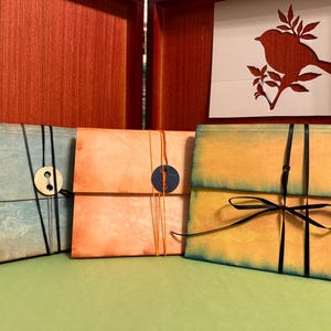 May include: Three handmade paper envelopes in shades of blue, peach, and yellow, each secured with a contrasting string or ribbon. A wooden button and a navy blue circle accent the closures. A bird silhouette decorates the background.