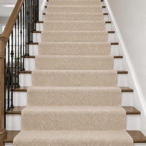 May include: A beige stair runner with a woven herringbone pattern covering a set of wooden stairs. The runner features a textured surface and runs vertically up the center of the staircase, which is flanked by a dark wood handrail and white risers.