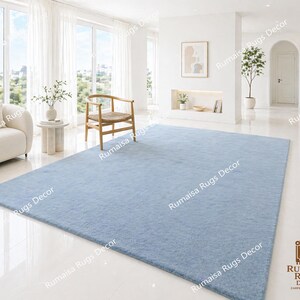 May include: A large, light blue rectangular rug dominates a bright, airy living room. The rug is placed on a glossy white floor, with a cream-colored sofa and wooden chair visible. The room features large windows and white curtains, creating a minimalist aesthetic.
