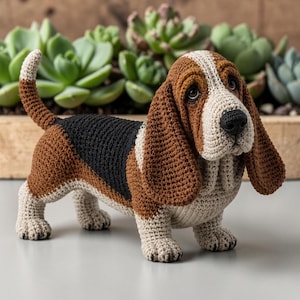 May include: A crocheted Basset Hound figurine featuring a tricolor pattern of brown, black, and cream yarn. The dog has long, drooping ears, a black nose, and dark eyes, standing in a classic pose with its tail held upright and alert.