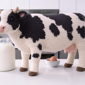May include: A handcrafted crochet cow with black and white spots, brown hooves, and small horns. A glass jar of milk and a small bowl with an egg are next to the cow. The cow is a decorative item.