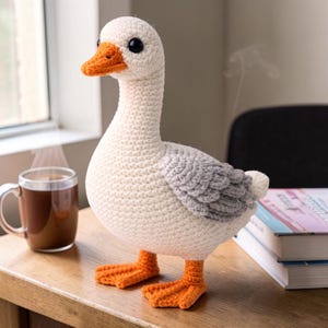 Könnte beinhalten: Ein handgefertigtes Häkelgans-Spielzeug mit weißem Körper, grauen Flügeln und orangefarbenem Schnabel und Füßen. Die Gans steht auf einer Holzoberfläche. Eine Tasse Kaffee und Bücher befinden sich im Hintergrund.