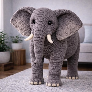 May include: A handmade, grey crochet elephant with large, floppy ears and ivory tusks. The amigurumi elephant has big, black eyes and is standing on a grey rug. The elephant is a decorative item.