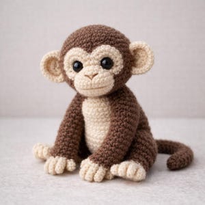 以下が含まれることがあります： 茶色とクリーム色の手作りの編み物モンキー。 ぬいぐるみは丸い頭、大きな黒い目、笑顔の顔をしています。 モンキーは腕と脚を伸ばし、丸まった尻尾で座っています。