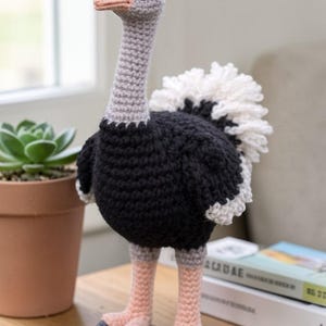 May include: A handmade crochet ostrich figurine. The bird has a gray head and neck, a black body, and pink legs and feet. The tail is white. The ostrich is standing on a wooden surface next to a potted plant.