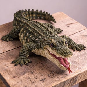 May include: A handcrafted, crochet alligator toy in shades of green and tan, with a detailed textured body, open mouth, and a pink tongue. The alligator is lying on a wooden surface, showcasing its realistic features and craftsmanship.