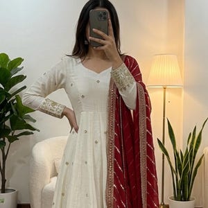 May include: A long, white dress with gold embroidery on the sleeves and hem. A red scarf with gold accents is draped over the shoulder. The dress has a V-neck and a flowing skirt. The brand name "NARI Outfits" is visible at the bottom of the dress.