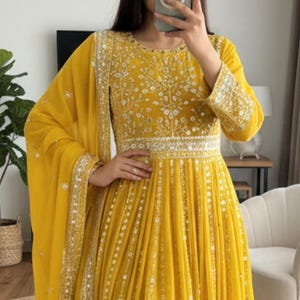May include: A long, yellow dress with intricate embroidery and embellishments. The dress features a fitted bodice, a flowing skirt, and long sleeves. The image also shows a matching yellow scarf. The brand name "NARI FASHIONINDIA" is visible.