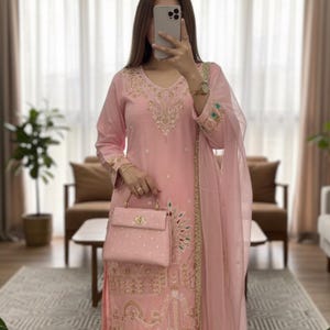 Puede incluir: Un traje salwar kameez rosa claro bordado con dupatta y bolso a juego. El traje presenta intrincados bordados dorados y coloridos en el escote, las mangas y el dobladillo. El bolso de mano también es rosa claro con un cierre dorado.