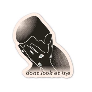 May include: A sticker featuring a black and white illustration of a person covering their face with their hands. The text "dont look at me" is printed in a simple font at the bottom of the sticker. The sticker has a light beige border.