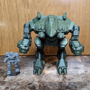 May include: A large olive green robotic action figure with a pointed head, articulated limbs, and clawed feet, positioned next to a smaller, unpainted gray robotic action figure standing on a black hexagonal base, both featuring detailed mechanical armor.